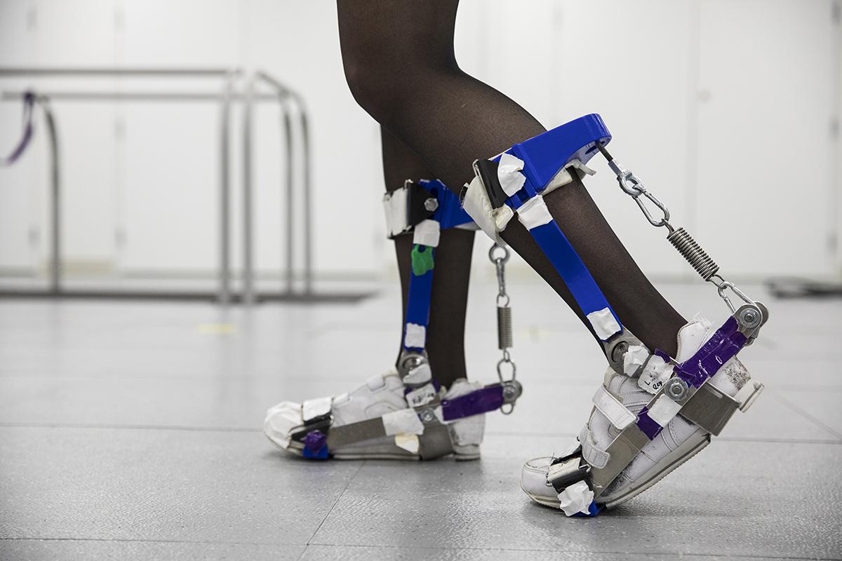 A woman walks across a lab wearing braces on both legs, from foot to calf. 